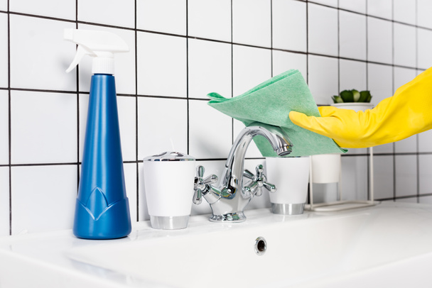 Vista ritagliata della persona nel rubinetto di pulizia del guanto di gomma con straccio vicino alla bottiglia di detergente sul lavandino in bagno  - Foto, immagini