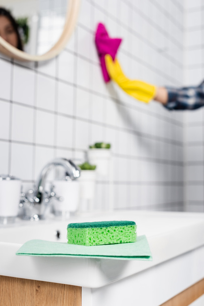 Σφουγγάρι και κουρέλι στο νεροχύτη κοντά γυναίκα καθαρισμού πλακιδίων στο μπάνιο σε θολή φόντο  - Φωτογραφία, εικόνα