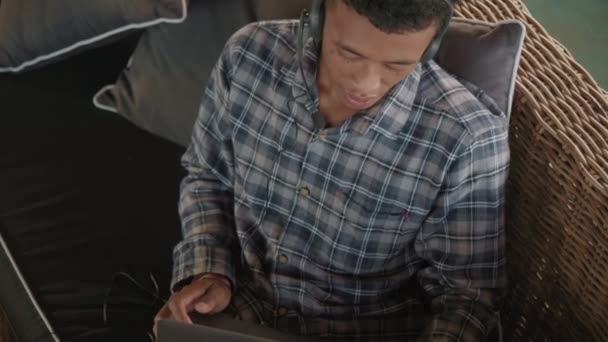 Vista de ángulo alto del joven asiático con camisa a cuadros, pantalones cortos y auriculares sentados en el sofá en la terraza con el ordenador portátil en las rodillas y consultando al cliente en línea - Metraje, vídeo