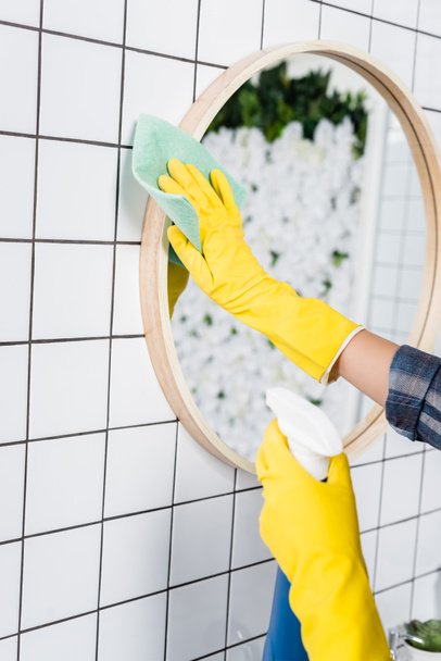 Cropped view of woman in yellow rubber gloves cleaning mirror with detergent and rag in bathroom  - Foto, Bild