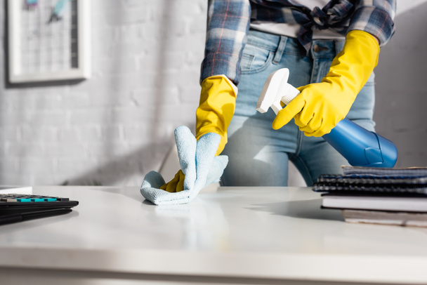 Vue recadrée de la femme en gants de caoutchouc table de nettoyage avec chiffon et détergent près de la calculatrice sur le premier plan flou  - Photo, image
