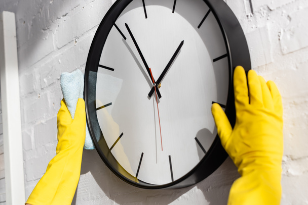 Cropped view of person in rubber gloves cleaning clock with rag on wall at home  - Photo, Image