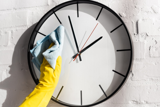 Vue recadrée de la personne dans un gant en caoutchouc horloge de nettoyage avec chiffon sur mur de briques blanches  - Photo, image