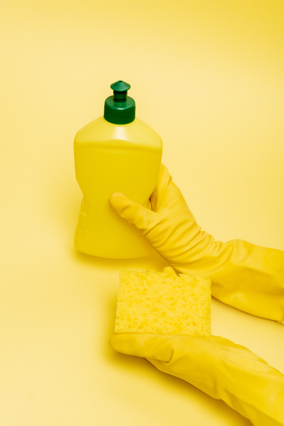 Vista en ángulo alto de las manos con guantes de goma que sujetan la esponja y el líquido lavavajillas sobre fondo amarillo - Foto, Imagen