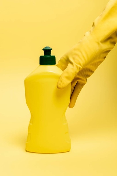 Vue recadrée de la main dans un gant en caoutchouc prenant du liquide vaisselle sur fond jaune - Photo, image