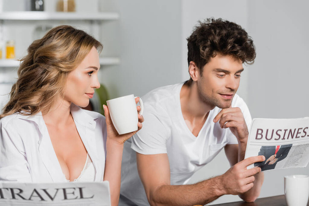 Сексуальная женщина с чашкой глядя на парня читает деловую газету на кухне  - Фото, изображение