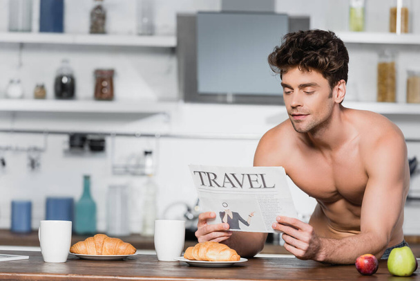 Sexy man reading travel newspaper near croissants and coffee in kitchen  - 写真・画像