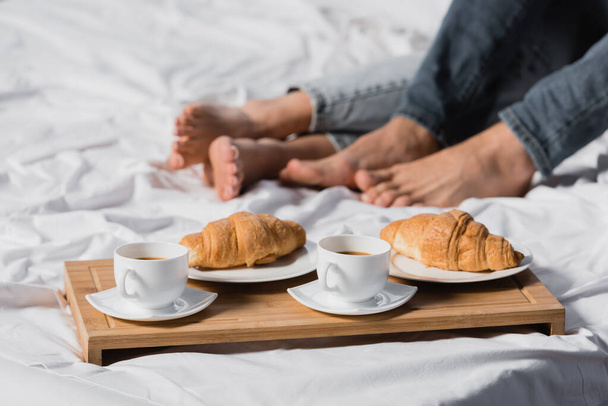 Copped view of coffee and croissants on breakfast tray near legs of couple on blurred background on bed  - Fotoğraf, Görsel