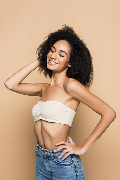 cheerful african american woman with bare shoulders in top and jeans standing with hand on hip on beige  - Foto, Imagem