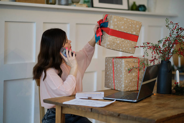 Pretty young lady sitting at desk and typing on laptop and talking on the phone with present boxes icons. box receive good online shop purchase at home, post mail shipping delivery concept - Photo, Image