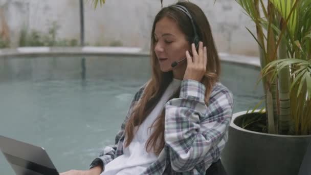 Mediana toma de la joven mujer caucásica con ropa casual y auriculares sentados cerca de la piscina con el ordenador portátil en las rodillas y el cliente de consulta por teléfono - Metraje, vídeo