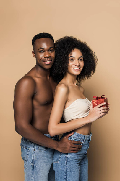 shirtless african american man hugging happy woman with present on beige - Фото, зображення
