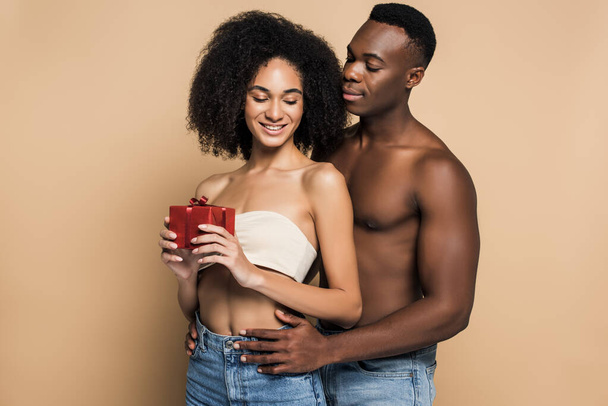 shirtless african american man hugging cheerful woman with present on beige - Photo, Image