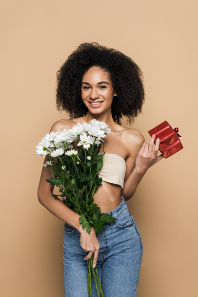 happy african american woman with bare shoulders holding present and flowers on beige - Valokuva, kuva