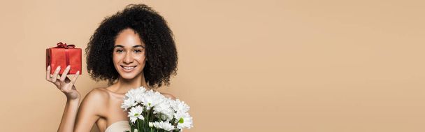 mulher americana africana alegre com ombros nus segurando presente e flores isoladas em bege, bandeira - Foto, Imagem