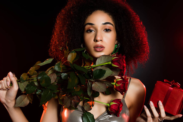 curly african american woman holding gift box and roses isolated on black  - Φωτογραφία, εικόνα