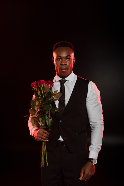 stylish african american man in formal wear with vest holding red roses on black  - Fotografie, Obrázek