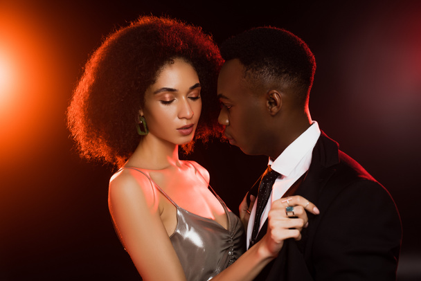 curly african american woman in dress undressing boyfriend in suit on black - 写真・画像