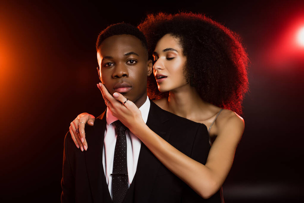 curly african american woman touching face of boyfriend in suit on black - Foto, afbeelding