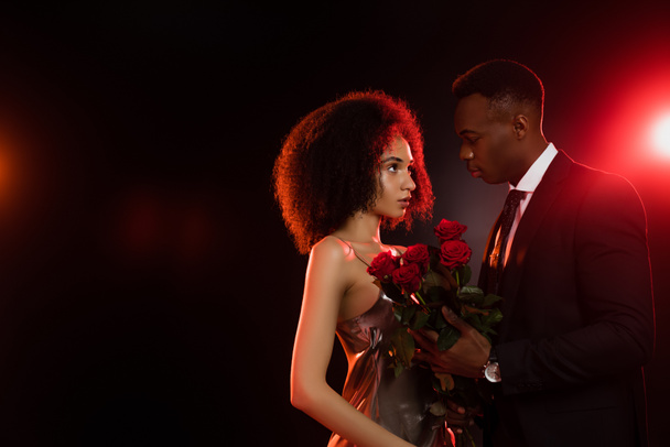 curly african american woman with red roses looking at boyfriend in suit on black - Фото, изображение