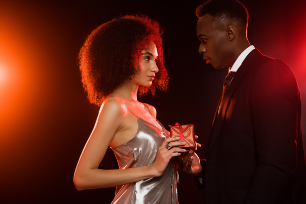 side view of african american man giving present to woman in dress on black - Foto, imagen