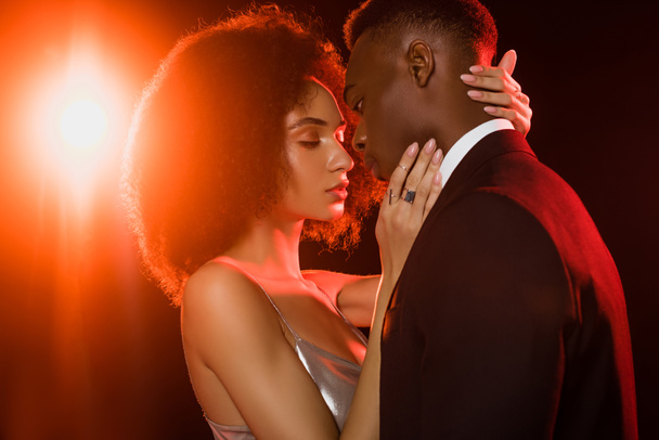 curly african american woman in dress embracing man on black - Zdjęcie, obraz