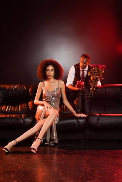 African american man holding red roses and gift box near elegant woman sitting on sofa and black background  - Foto, afbeelding