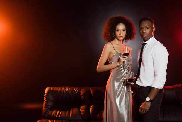 curly african american woman and man holding glasses of red wine on black  - Foto, afbeelding