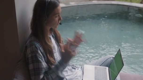 Άποψη υψηλής γωνίας της νεαρής Καυκάσιας γυναίκας που φοράει casual πουκάμισο, τζιν και ακουστικά κάθεται στον καναπέ κοντά στην πισίνα με το laptop στα γόνατα και συμβουλεύεται τον πελάτη τηλεφωνικά - Πλάνα, βίντεο