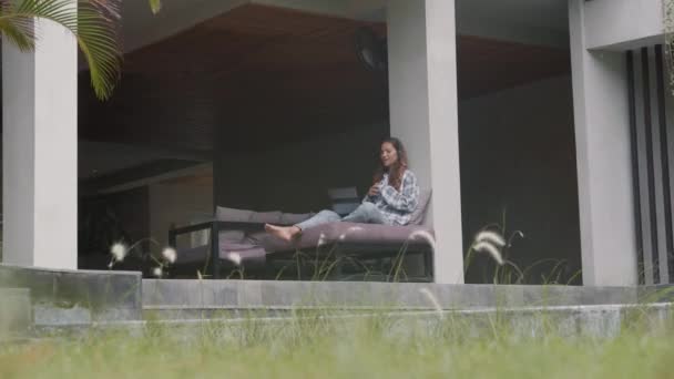 Široký záběr mladé dlouhosrsté ženy v ležérní košili, džínách a sluchátkách sedí na pohovce na terase v moderním domě s notebookem na kolenou a mají video setkání s kolegou - Záběry, video