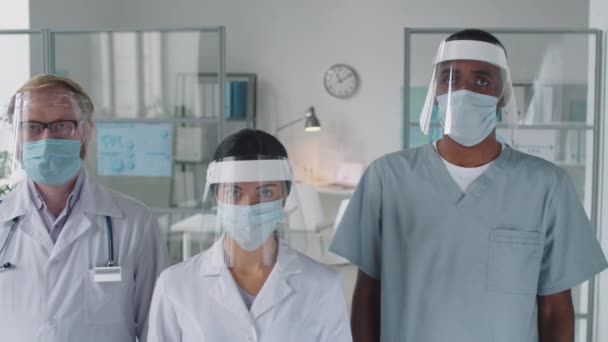 Ritratto di squadra di medici multietnici in uniforme, maschere protettive e schermi in piedi insieme nello studio medico e in posa per la fotocamera mentre si lavora durante la pandemia covid-19 - Filmati, video