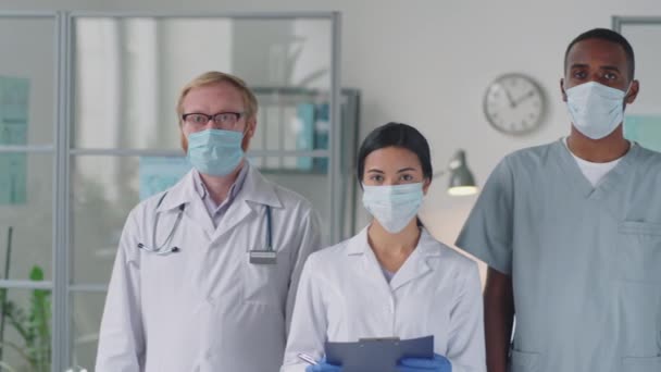 Portrait of team of diverse doctors in professional uniform and protective masks standing together in medical office and posing for camera while working during coronavirus outbreak - Footage, Video