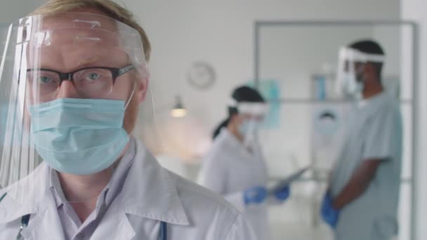 Chest up sledování záběru mužského lékaře v ochranném obličejovém skle, laboratorním plášti a lékařské masce při pohledu na kameru při práci v nemocnici během pandemie koronaviru - Záběry, video