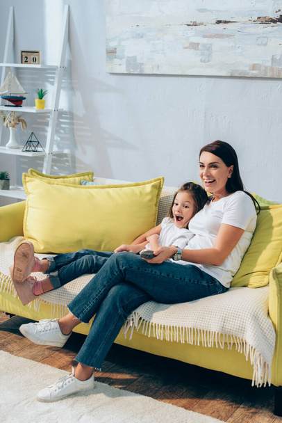 Volle Länge der glücklichen Mutter mit Fernbedienung, die aufgeregte Tochter auf der Couch umarmt - Foto, Bild