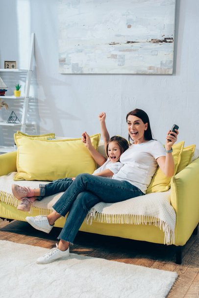 Full length of excited mother with remote controller and daughter looking away on couch at home - Photo, Image
