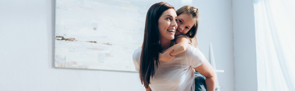 ευτυχισμένη μητέρα κοιτάζοντας μακριά, ενώ piggybacking κόρη στο σπίτι σε θολή φόντο, πανό - Φωτογραφία, εικόνα