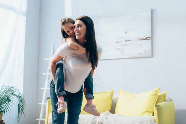 happy mother looking away while piggybacking daughter in living room on blurred background, banner - Foto, Bild