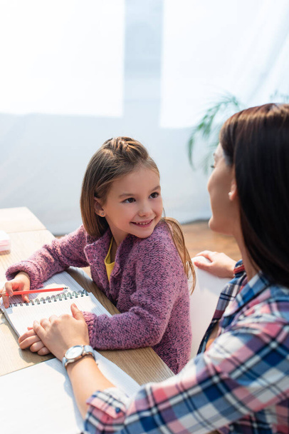 Счастливая дочь с карандашом и блокнотом смотрит на мать, сидя за столом на размытом фоне - Фото, изображение