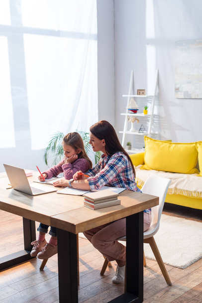 Full length of smiling mother sitting near daughter writing in notebook near laptop in living room - Photo, Image