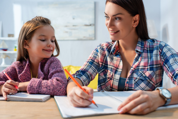 Usmívající se matka při pohledu na dceru při psaní v kopii knihy na stole na rozmazaném pozadí - Fotografie, Obrázek