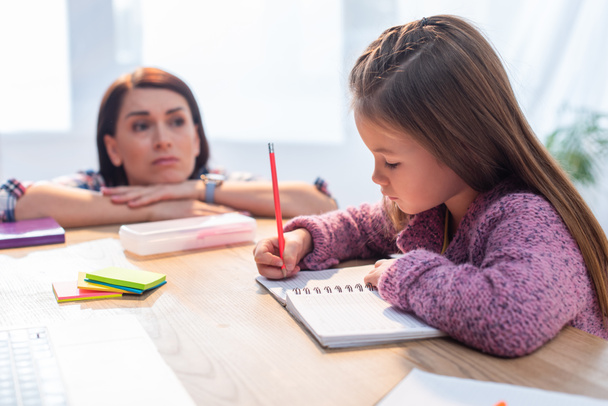 Upset mother looking at daughter writing in notebook at desk on blurred background - Photo, Image