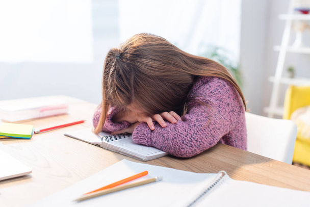 Exhausted girl leaning on hands at desk near stationery on blurred background - Foto, imagen