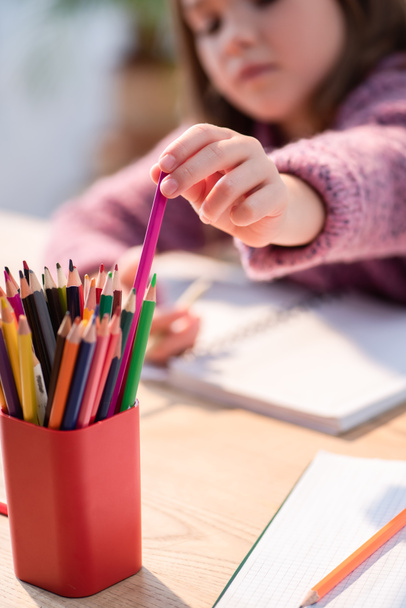 Oříznutý pohled na dívku, která bere barevnou tužku z držáku na stole na rozmazaném pozadí - Fotografie, Obrázek