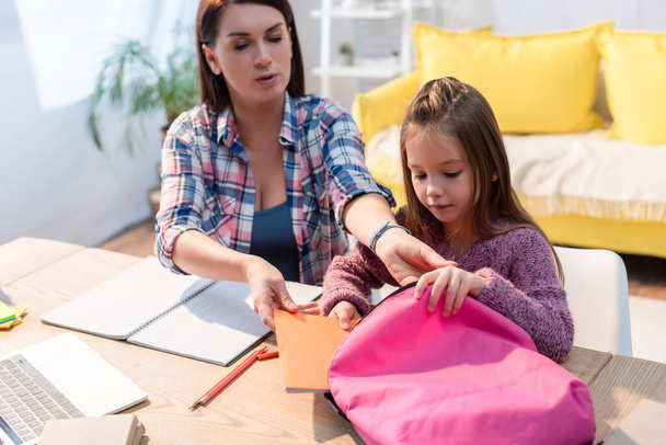 Mother and daughter putting copy book in backpack on desk at home on blurred background - Фото, изображение