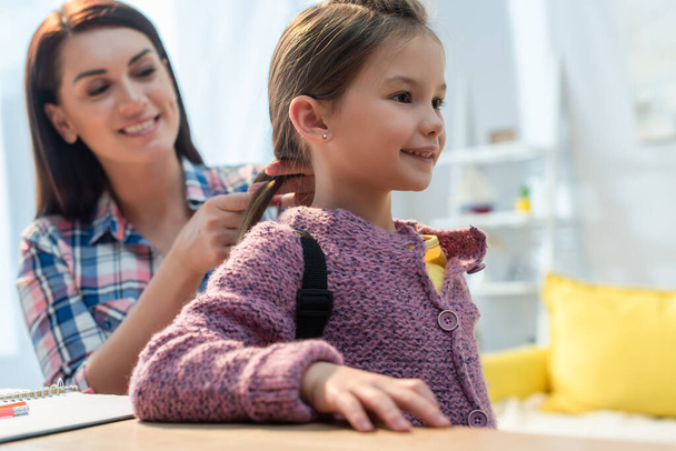 Happy mother plaiting hair of daughter with backpack at home on blurred background - Φωτογραφία, εικόνα
