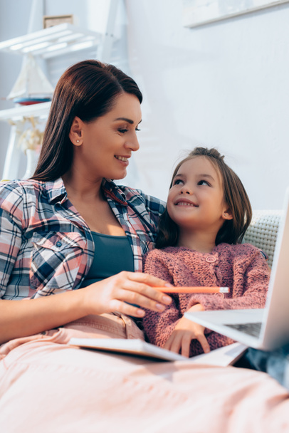 Smiling mother looking at daughter while pointing with pencil at blurred laptop on foreground - Фото, изображение