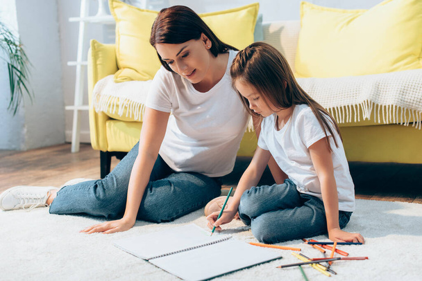 Mother sitting near daughter drawing in copy book on floor on blurred background - Foto, Imagem