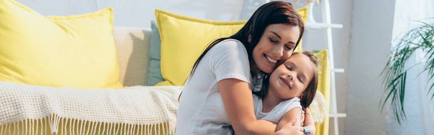 Onnellinen äiti suljetuin silmin halaamalla tytärtä lähellä sohvaa kotona, banneri - Valokuva, kuva