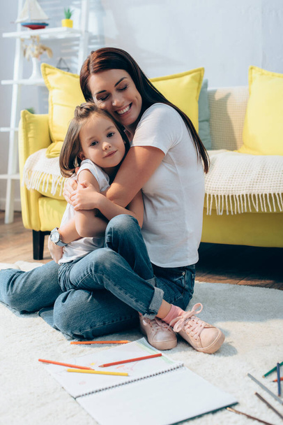 Cheerful mother with closed eyes hugging daughter while sitting on floor on blurred background - Foto, Bild