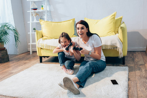 KYIV, UKRAINE - OCTOBER 19, 2020: Full length of excited mother and daughter with open mouths playing with gamepads on floor at home - Foto, Imagem